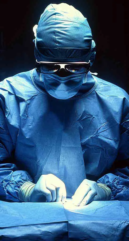 Chirurg in blauer OP-Kleidung mit Maske und Brille, arbeitet konzentriert unter OP-Licht